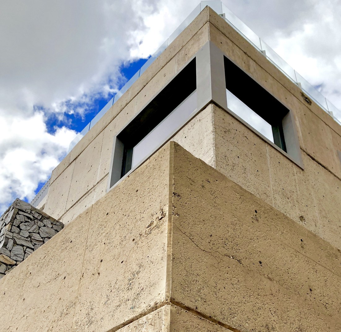 Corner of a rammed-earth wall seen from below: the lifts, the roof parapet and the glass rail against the sky